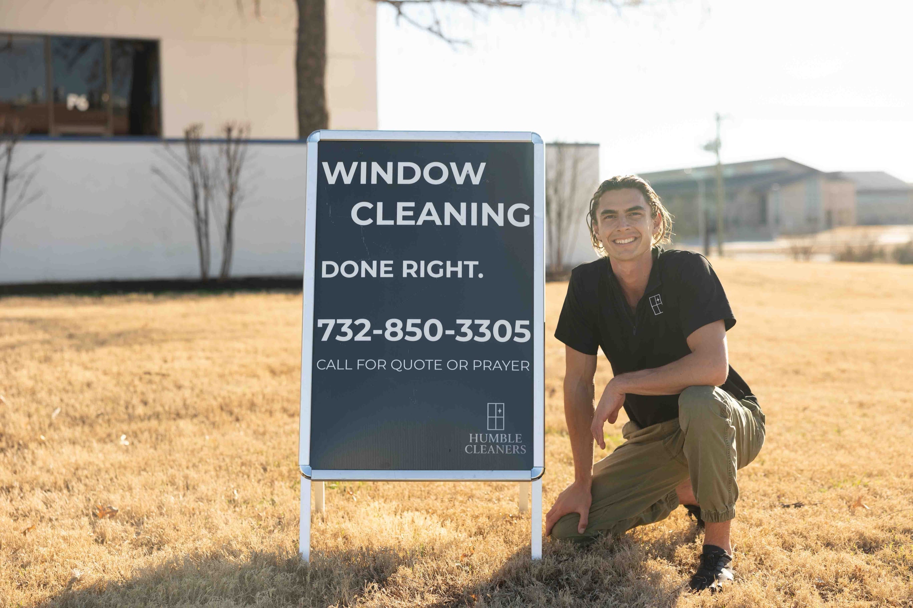 A crew member kneeling beside the Humble Cleaners yard sign, which reads: window cleaning done right — call for quote or prayer